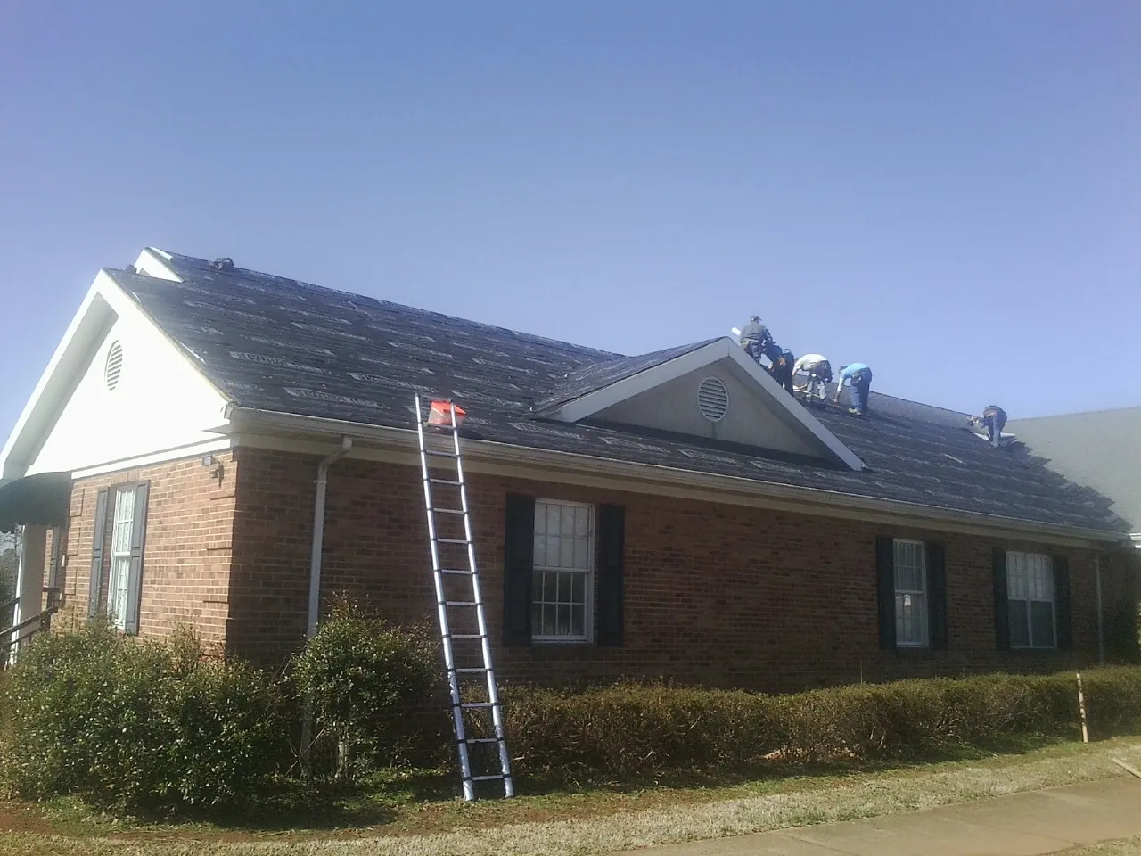 Crew tearing shingles off a brick ranch roof with a ladder set — Emergency Roof Repair work by our crew in Saint Simons Island