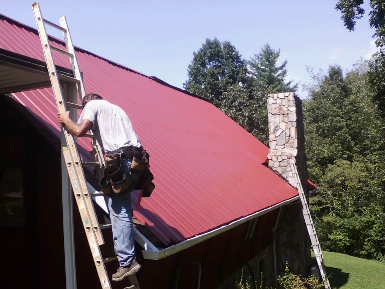 Roofer on a ladder beside a red metal roof and stone chimney — Soffit & Fascia Repair by Roofing Brothers in Saint Simons Island